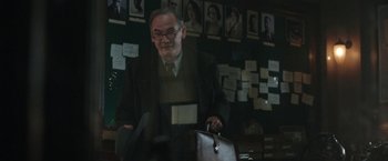 Movie still from “Their Finest” (2016), directed by Lone Scherfig – An older man holding a briefcase in front of a bunch of pictures; Medium shot, Over the shoulder angle
