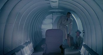 Movie still from “This Island Earth” (1955), directed by Joseph M. Newman – A man in a suit and tie standing in an airplane; Wide shot, Overhead angle