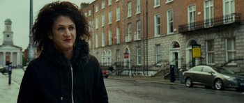 Movie still from “This Must Be the Place” (2011), directed by Paolo Sorrentino – A woman is walking down the street wearing a black jacket; Medium shot, Over the shoulder angle