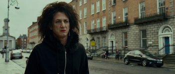 Movie still from “This Must Be the Place” (2011), directed by Paolo Sorrentino – A person with long black hair standing on the side of the street; Close Up shot, Over the shoulder angle