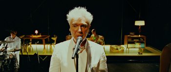 Movie still from “This Must Be the Place” (2011), directed by Paolo Sorrentino – A man in a white suit is holding a microphone; Close Up shot, Over the shoulder angle