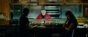 Movie still from “This Must Be the Place” (2011), directed by Paolo Sorrentino – A person sitting at a table in front of a counter; Medium shot, Over the shoulder angle