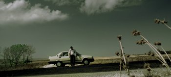 Movie still from “Three Monkeys” (2008), directed by Nuri Bilge Ceylan – A man standing next to a white car on the side of the road; Wide shot, Low angle