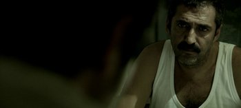 Movie still from “Three Monkeys” (2008), directed by Nuri Bilge Ceylan – A man in a white tank top is sitting in a dark room; Close Up shot, Over the shoulder angle