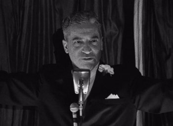 An older man in a black suit is holding a microphone in front of him