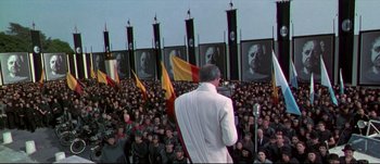 Movie still from “Titus” (1999), directed by Julie Taymor – A man in a white suit is giving a speech to a crowd of people; Extreme Wide shot, High angle