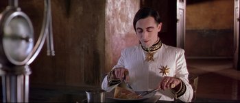 Movie still from “Titus” (1999), directed by Julie Taymor – A man in a white shirt is cutting a piece of pizza; Medium shot, Low angle