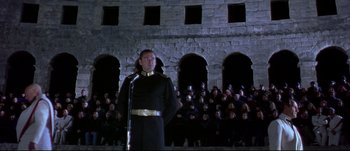Movie still from “Titus” (1999), directed by Julie Taymor – A man in a black suit stands in front of a group of people; Wide shot, Low angle
