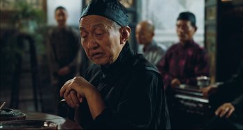 Movie still from “To Live” (1994), directed by Yimou Zhang – An older man in a black hat is praying; Medium shot, Over the shoulder angle