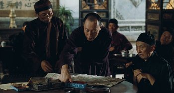 Movie still from “To Live” (1994), directed by Yimou Zhang – A man in a monk's outfit is cutting paper; Medium shot, Over the shoulder angle