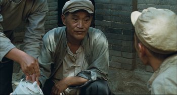 Movie still from “To Live” (1994), directed by Yimou Zhang – A man sitting on the ground in front of a brick wall; Medium shot, High angle