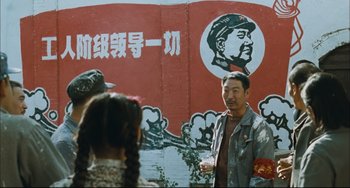 Movie still from “To Live” (1994), directed by Yimou Zhang – A man standing in front of a mural of mao zedong; Medium shot, Over the shoulder angle