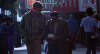 Movie still from “To Live and Die in L.A.” (1985), directed by William Friedkin – Two men walking down the street while one of them is holding a box; Medium shot, Low angle