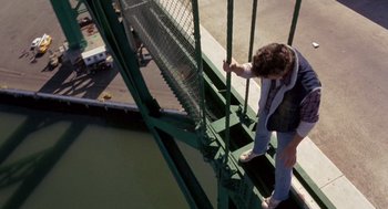 Movie still from “To Live and Die in L.A.” (1985), directed by William Friedkin – A man standing on a metal railing near a body of water; Wide shot, Overhead angle
