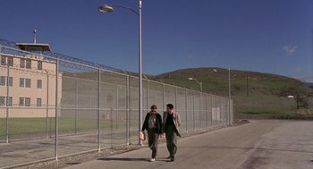 Movie still from “To Live and Die in L.A.” (1985), directed by William Friedkin – Two men walking down a street near a fence; Wide shot, Low angle