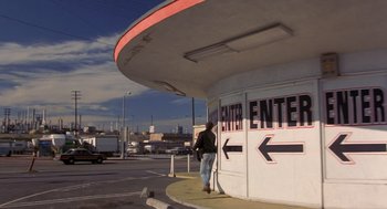 Movie still from “To Live and Die in L.A.” (1985), directed by William Friedkin – A woman standing outside of a building with a sign pointing to the center entrance; Wide shot, Low angle