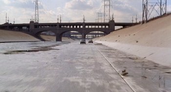 Movie still from “To Live and Die in L.A.” (1985), directed by William Friedkin – Two cars driving down a road with a bridge in the background; Extreme Wide shot, High angle