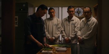 Movie still from “Tokyo Vice” (2022), created by J.T. Rogers – A group of men standing around a table cutting vegetables; Medium shot, Over the shoulder angle