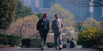 Movie still from “Tokyo Vice” (2022), created by J.T. Rogers – Two businessmen walking down a sidewalk near a park; Wide shot, Over the shoulder angle