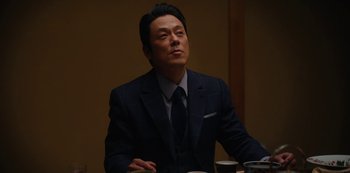 Movie still from “Tokyo Vice” (2022), created by J.T. Rogers – A man wearing a suit and tie sitting at a table; Medium shot, Low angle
