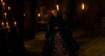 Movie still from “Tom Thumb” (2001), directed by Olivier Dahan – A woman in a black and gold dress standing next to lit torches; Wide shot, Over the shoulder angle