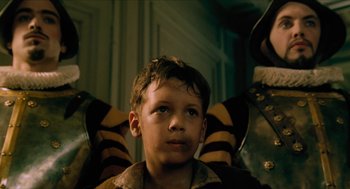 Movie still from “Tom Thumb” (2001), directed by Olivier Dahan – A young boy wearing a brown jacket and a brown and yellow striped jacket; Close Up shot, Low angle