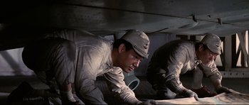 Movie still from “Tora! Tora! Tora!” (1970), directed by Kinji Fukasaku – A man wearing a hat is working under a vehicle; Medium shot, Low angle