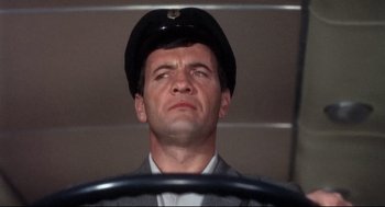 Movie still from “Torn Curtain” (1966), directed by Alfred Hitchcock – A man wearing a hat and a suit is sitting behind a steering wheel; Close Up shot, Low angle