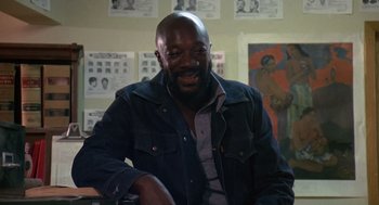 Movie still from “Truck Turner” (1974), directed by Jonathan Kaplan – A man sitting in front of a wall with pictures on the wall behind him; Medium shot, High angle