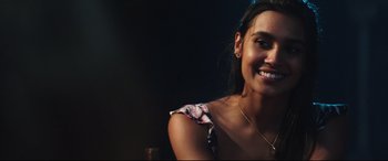 Movie still from “Truth or Dare” (2018), directed by Jeff Wadlow – A woman smiles brightly while sitting in front of a dark background; Close Up shot, Over the shoulder angle