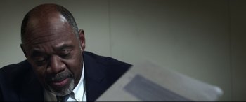 Movie still from “Truth or Dare” (2018), directed by Jeff Wadlow – A man in a suit and tie sitting in front of papers; Close Up shot, Low angle