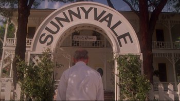 Movie still from “Twilight Zone: The Movie” (1983), directed by Steven Spielberg – A man standing in front of a white building with a sign on it; Wide shot, Low angle