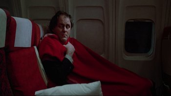 Movie still from “Twilight Zone: The Movie” (1983), directed by Steven Spielberg – A man wrapped in a blanket sitting on a plane; Medium shot, Low angle