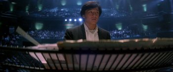 Movie still from “Twin Dragons” (1992), directed by Ringo Lam – A man in a black jacket is sitting in front of an audience; Medium shot, Low angle