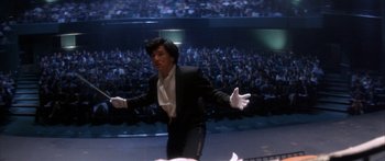 Movie still from “Twin Dragons” (1992), directed by Ringo Lam – A man standing in front of an audience wearing a suit and white gloves; Medium shot, Low angle