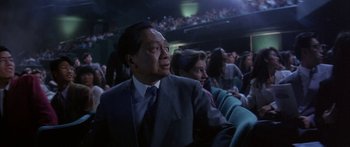 Movie still from “Twin Dragons” (1992), directed by Ringo Lam – A man in a suit and tie sitting in front of an audience; Medium shot, Low angle