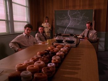 Movie still from “Twin Peaks” (1990), created by David Lynch – A group of people sitting around a table with donuts on top of it; Wide shot, High angle
