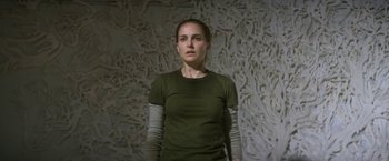 Movie still from “Annihilation” (2018), directed by Alex Garland – A woman in a green shirt and arm warmers stands in front of a wall; Medium shot, Overhead angle