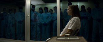 Movie still from “Annihilation” (2018), directed by Alex Garland – A group of people wearing masks in a room with a woman sitting at a table; Medium shot, Over the shoulder angle