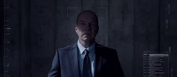 Movie still from “Anon” (2018), directed by Andrew Niccol – A man wearing a suit and tie in front of a concrete wall; Medium shot, Over the shoulder angle
