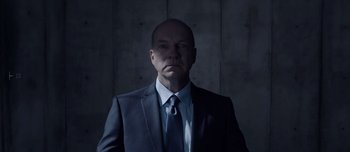 Movie still from “Anon” (2018), directed by Andrew Niccol – A man wearing a suit and tie in a dark room; Medium shot, Low angle