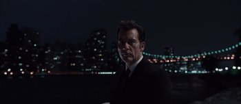 Movie still from “Anon” (2018), directed by Andrew Niccol – A man wearing a suit and tie standing in front of a city skyline at night; Medium shot, Low angle