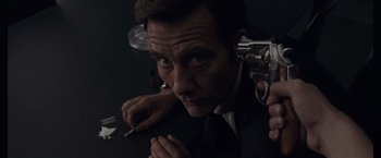 Movie still from “Anon” (2018), directed by Andrew Niccol – A man in a black suit smoking a cigarette and holding a gun in his hand; Close Up shot, High angle