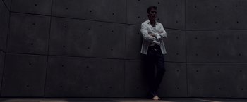 Movie still from “Anon” (2018), directed by Andrew Niccol – A man standing next to a wall with his feet crossed; Medium shot, Low angle