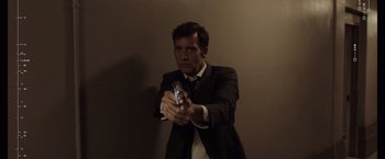 Movie still from “Anon” (2018), directed by Andrew Niccol – A man in a suit and tie holding a gun in his hand; Medium shot, Low angle