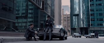 Movie still from “Anon” (2018), directed by Andrew Niccol – Two men sitting on the back of a car on the side of the road; Wide shot, High angle