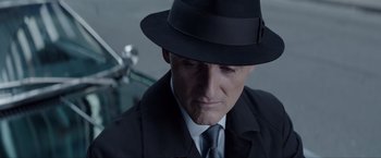 Movie still from “Anon” (2018), directed by Andrew Niccol – A person wearing a suit and a hat; Close Up shot, Over the shoulder angle