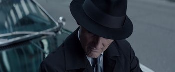 Movie still from “Anon” (2018), directed by Andrew Niccol – A man wearing a black hat and a black coat; Close Up shot, Over the shoulder angle
