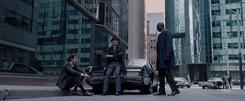 Movie still from “Anon” (2018), directed by Andrew Niccol – A group of men standing around a car on a city street; Wide shot, High angle