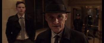 Movie still from “Anon” (2018), directed by Andrew Niccol – An older man wearing a black hat and a black suit; Close Up shot, Over the shoulder angle